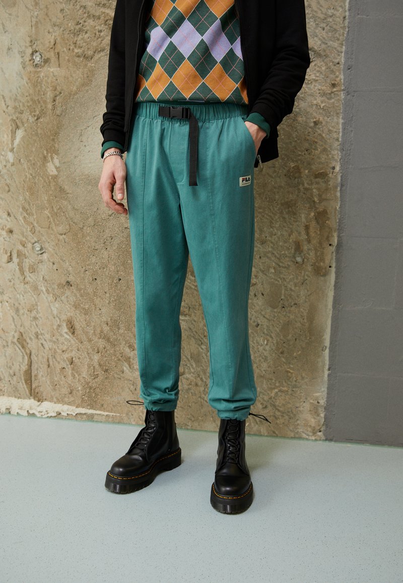 Teal joggebukser med svart belte; har Fila-logo. Brukes med en mønstret genser og svarte snørebånds plattformsko.