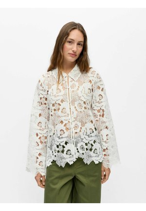 Femme portant un chemisier en dentelle florale blanche à manches longues avec un col et un pantalon large taille haute vert olive sur fond blanc.