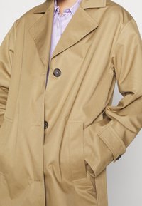 Trench-coat beige avec col à revers, deux boutons visibles, poches latérales et lanières aux poignets, porté sur une chemise violette claire à col.