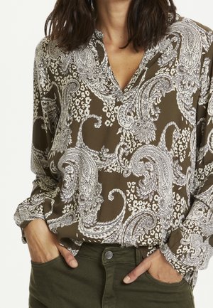 Kvinde iført en olivengrøn bluse med paisleymønster og lange ærmer samt mørkegrønne bukser, hænderne stukket i forlommerne.