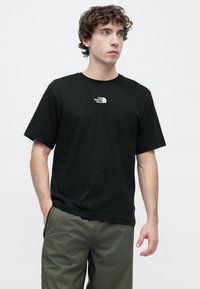 Μαύρο βαμβακερό μπλουζάκι με μικρό λευκό λογότυπο στο στήθος, κοντά μανίκια και λαιμόκοψη τύπου crew. Το ύφασμα φαίνεται απαλό και λείο.