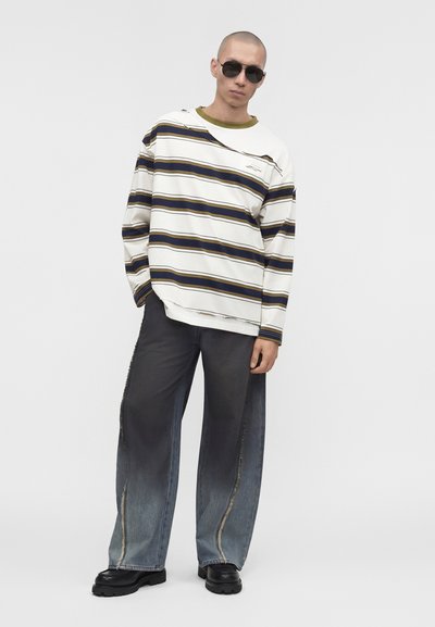 Άνδρας που φοράει μαύρα γυαλιά ηλίου, υπερμεγέθη ριγέ πουλόβερ, φαρδιά ξεβαμμένα μαύρα και μπλε τζιν και μαύρα χοντροκομμένα παπούτσια, στέκεται μπροστά σε λευκό φόντο.