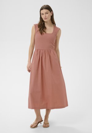 Kobieta o długich brązowych włosach, ubrana w bezrękawnikową sukienkę midi w kolorze przygaszonego różu oraz beżowe sandały na obcasie, stojąca na jednolitym tle.