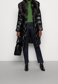 Abrigo acolchado largo negro con cuello de piel, combinado con un suéter de cuello alto verde, jeans oscuros y botines negros, llevando un bolso negro.