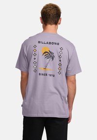 Fialové bavlněné tričko s grafikou palmy a slunce na zadní straně, s textem "BILLABONG SINCE 1973" a dekorativními prvky.