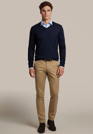 Jonge man draagt een marineblauwe V-hals trui over een lichtblauw overhemd, beige broek en donkerbruine schoenen, staand tegen een effen achtergrond.