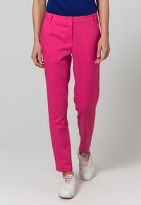 Kobieta ubrana w jaskraworóżowe spodnie o prostych nogawkach z guzikiem i zamkiem błyskawicznym, zestawione z białymi trampkami i granatową bluzką.