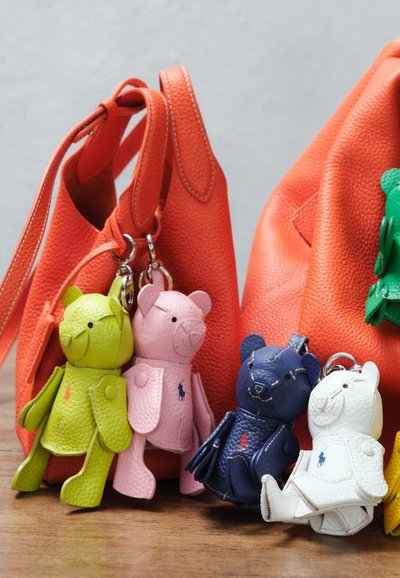 Vijf kleurrijke leren sleutelhangers in de vorm van een beer in groen, roze, blauw, wit en geel, bevestigd aan of dicht bij twee oranje leren tassen op een houten oppervlak.