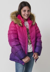 Chica sonriente que lleva una chaqueta de invierno con degradado en rosa y morado, con capucha de pelo, camiseta blanca gráfica y jeans azules, de pie frente a un fondo blanco.
