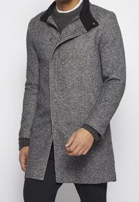 Grau melierter Wollmischmantel mit schwarzem Kragen, einreihigem Design, taillierter Passform und gerippten Strickbündchen. Auffällig durch sein strukturiertes Erscheinungsbild.