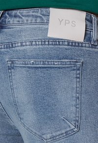 Světle modré džíny z denimu s bílou nášivkou "YPS". Zadní kapsa je viditelná s klasickým ševem a hladkým povrchem.