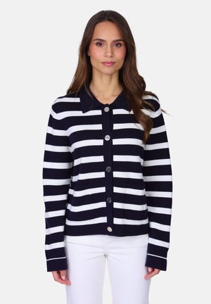 Femme portant un cardigan boutonné à rayures horizontales bleu marine et blanches avec des poches avant, et un pantalon blanc, debout devant un fond uni.