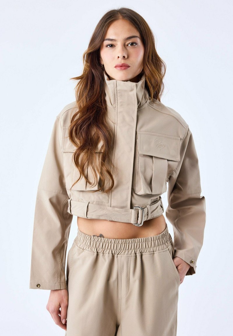 Beige cropped jasje met een hoge kraag, meerdere voorzakken en een ceintuursdetail. Gladde textuur met een subtiel geribbeld patroon.
