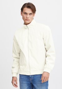 Hvid, letvægtsjakke med høj krave, frontlynlås og elastiske manchetter; parret med blå jeans. Glat tekstur og minimalistisk design.