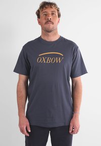 T-shirt en coton bleu marine à manches courtes et col rond, avec le texte doré "OXBOW" centré sur la poitrine. Design simple, texture lisse.
