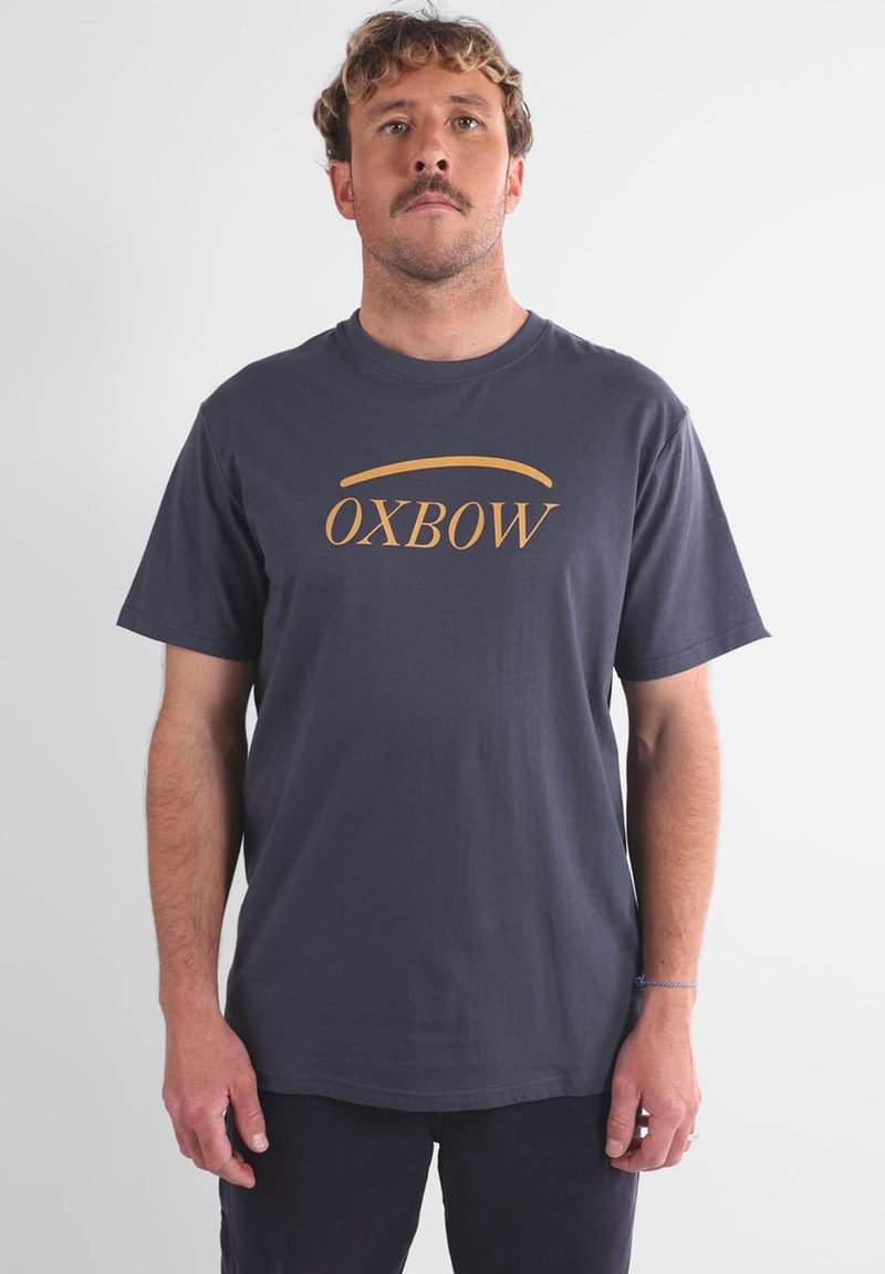 T-shirt en coton bleu marine à manches courtes et col rond, avec le texte doré "OXBOW" centré sur la poitrine. Design simple, texture lisse.