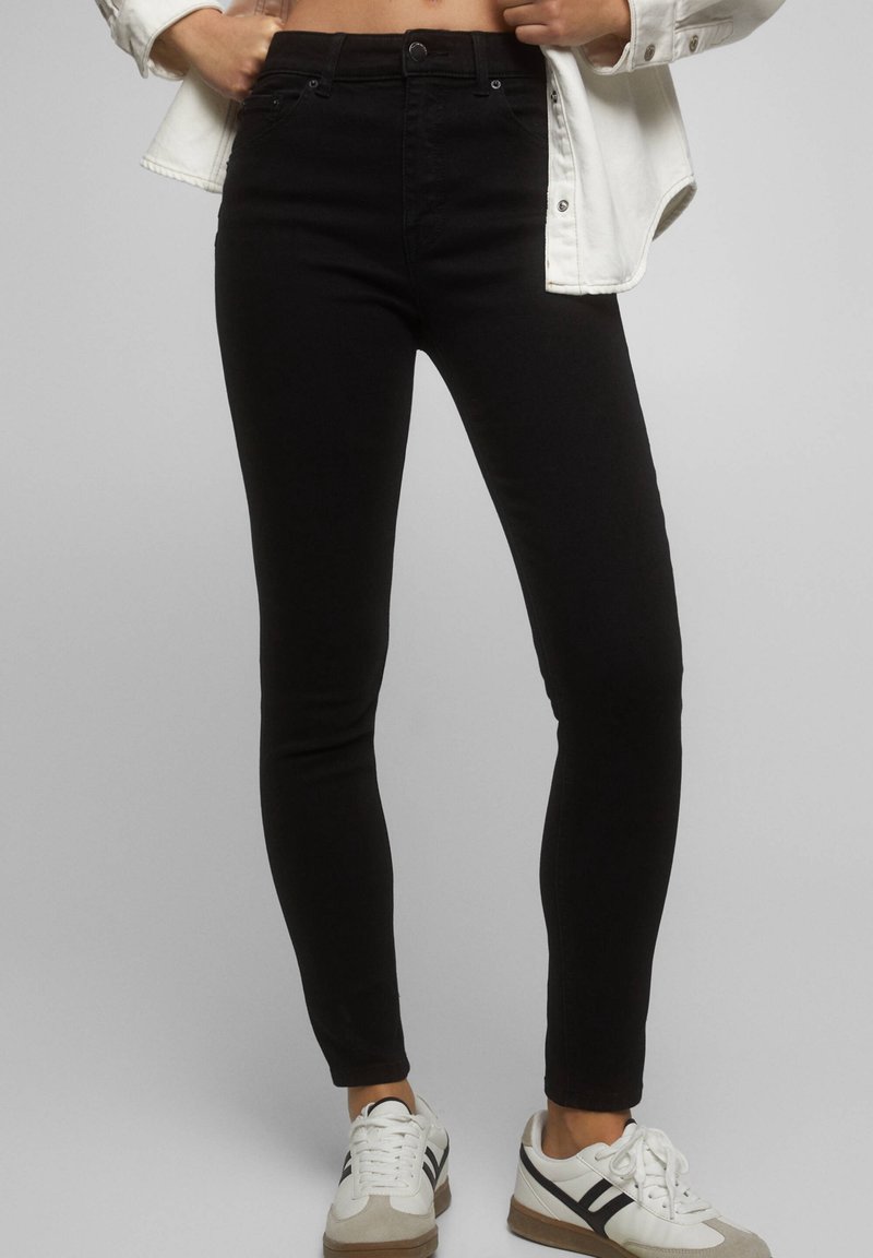 Personne portant un jean noir ajusté, des baskets blanches avec des bandes noires et une chemise blanche boutonnée tenant la taille.