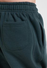 Dunkelblau-grüne Jogginghosen zeichnen sich durch eine weiche Textur, einen elastischen Bund und seitliche Taschen aus. Die Gesäßtasche hat eine rechteckige Form mit gesteppten Kanten.