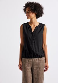 Blouse sans manches noire avec un décolleté en V, une taille élastique et une texture lisse, associée à un pantalon en lin léger de couleur neutre.