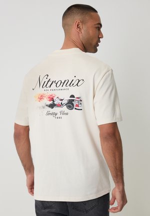 Mężczyzna w kremowej koszulce z napisem „Nitronix RZK Performance” i grafiką samochodu wyścigowego wydzielającego ogień i dym z tyłu.