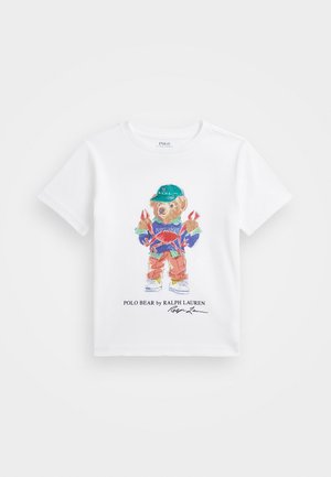 T-shirt blanc avec un ours coloré portant une casquette verte, une chemise rayée, un pantalon orange et tenant deux crabes rouges, avec le texte Ralph Lauren en dessous.