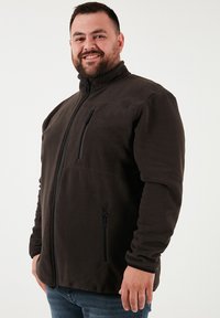 Barna polárdzseki magas gallérral, cipzáras mellszsebből és oldalsó zsebekből áll. Simább textúrával és laza szabással rendelkezik.