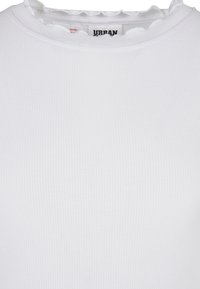 Detailní záběr na bílý žebrovaný top s volánkovým kulatým výstřihem a viditelnou vnitřní cedulkou.