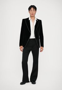 Sak hitam beludru dengan kerah satin, dipasangkan dengan kemeja putih tanpa kerah dan celana hitam. Model berdiri tegak di latar belakang netral.