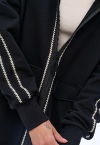 Czarny sweatshirt z zamkiem z przodu, z kremowymi zygzakowymi przeszyciami wzdłuż rękawów i kieszeni. Miękka tekstura i wykończone ściągaczem rękawy.