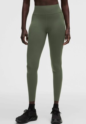 Persona che indossa leggings verde oliva a vita alta e scarpe da ginnastica nere, in piedi davanti a uno sfondo chiaro e uniforme.