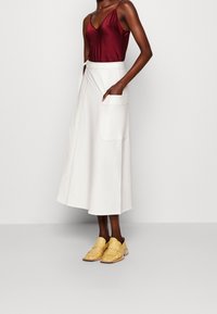 Witte midi-jurk met grote zak, hoge taille en subtiele stiksels, gecombineerd met een donkerrode satijnen cami en gele instappers.