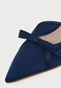Chaussure plate bleu marine à bout pointu avec un petit nœud en tissu sur le dessus et une doublure intérieure beige clair.