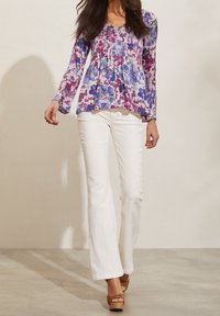 Blommig peplumtop i lila och blått, lång ärm, v-ringning, ihop med vita utsvängda byxor och bruna platåhälar på ljus bakgrund.