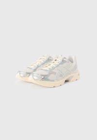 Sportowe trampki z białą siateczkową cholewką z srebrnymi akcentami, teksturowaną podeszwą i jasnobeżowymi sznurowadłami. Odbijające światło detale po bokach.