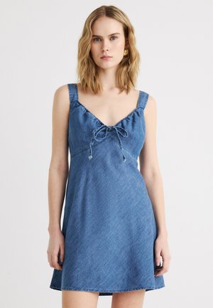 Νεαρή γυναίκα φορώντας μπλε μαν sleeveless φόρεμα από τζιν με λεπτές τιράντες και φιόγκο στη μέση, που στέκεται μπροστά σε απλό ανοιχτό φόντο.