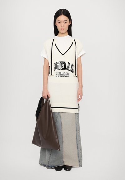 Knitted sleeveless sweater in cream with black accents and lettering, paired with a long, patchwork denim skirt. Model holds a brown tote bag.