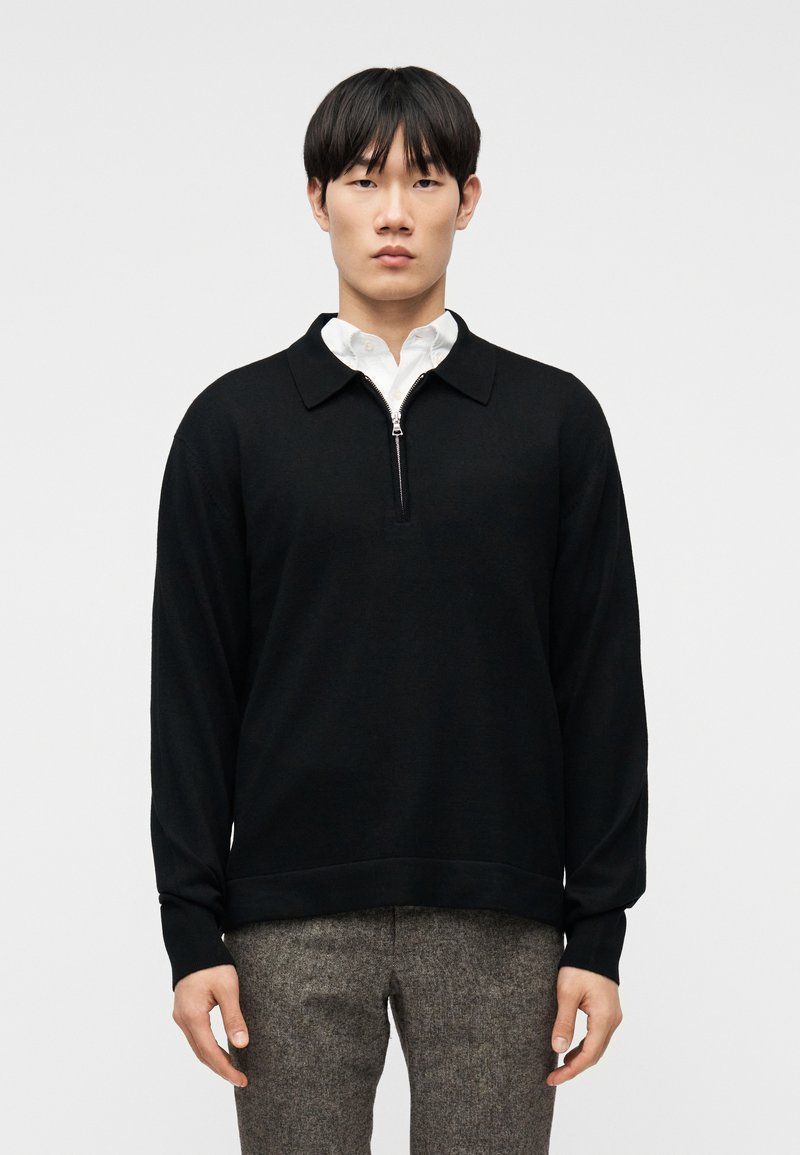 Άνδρας που φοράει μαύρο πουλόβερ μ ε φερμουάρ πάνω από λευκό πουκάμισο με γιακά και γκρι παντελόνι με σχέδια, στέκεται μπροστά σε απλό φωτεινό φόντο.