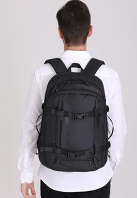 Zaino nero con spallacci regolabili, numerosi scomparti con zip e un tessuto testurizzato. Presenta dettagli in metallo e il logo.