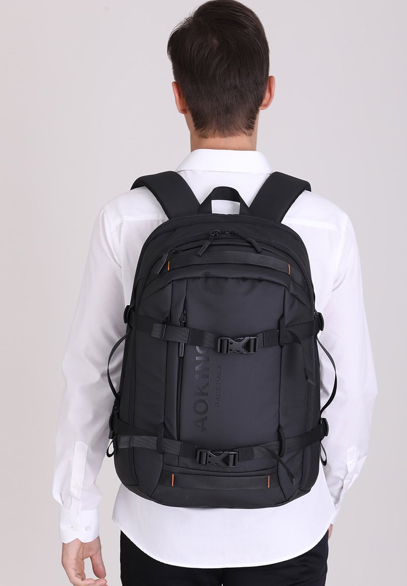 Zaino nero con spallacci regolabili, numerosi scomparti con zip e un tessuto testurizzato. Presenta dettagli in metallo e il logo.