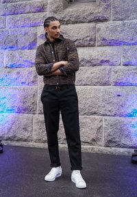 Camisa de botones marrón con estampado, pantalones negros ajustados con cordón y zapatillas blancas. El modelo está de pie contra una pared de piedra.