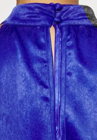 Top azul de satén com colar alto, apresentando dois botões redondos nas costas e um detalhe em fenda, mostrando uma textura lisa e brilhante.