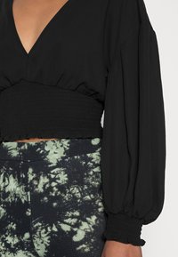 Top nero a maniche lunghe con scollatura a V profonda, corpetto arricciato e orlo a volants. Abbinato a pantaloni a motivi tie-dye verde scuro.