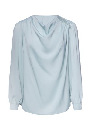 Lyseblå langærmet bluse med draperet halsudskæring og blomsterapplikation på den ene skulder, vist mod hvid baggrund.