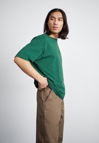 Grøn bomulds t-shirt med korte ærmer, afslappet pasform, der har et diskret logo. Sammen med brune, løstsiddende bukser med sidelommer.