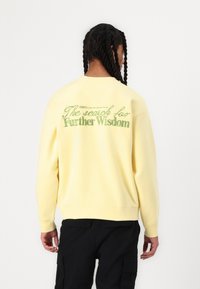 Yellow sweatshirt with a relaxed fit, featuring embroidered green text on the back that reads "The search for Further Wisdom."