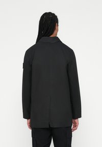 Cappotto nero con ampio collare, taglio dritto e maniche lunghe. Presenta uno spacco centrale posteriori e dettagli di cucitura sottili.