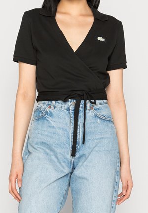 Vrouw draagt een zwart wikkelshirt met korte mouwen en een strikceintuur, gecombineerd met lichtblauwe jeans met hoge taille, voorzien van een klein krokodillenlogo op de borst.