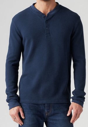 Man wearing a navy blue long-sleeve waffle-knit henley shirt with three buttons and dark blue jeans.