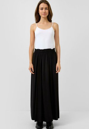 Junge Frau mit langen braunen Haaren, die ein weißes ärmelloses Oberteil und einen hoch taillierten schwarzen Maxirock trägt, steht barfuß vor einem schlichten Hintergrund.