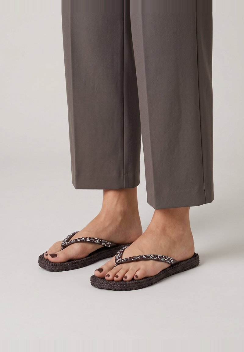 Pieds portant des tongs tissées foncées avec des brides florales, associées à un pantalon gris raccourci sur un fond uni.